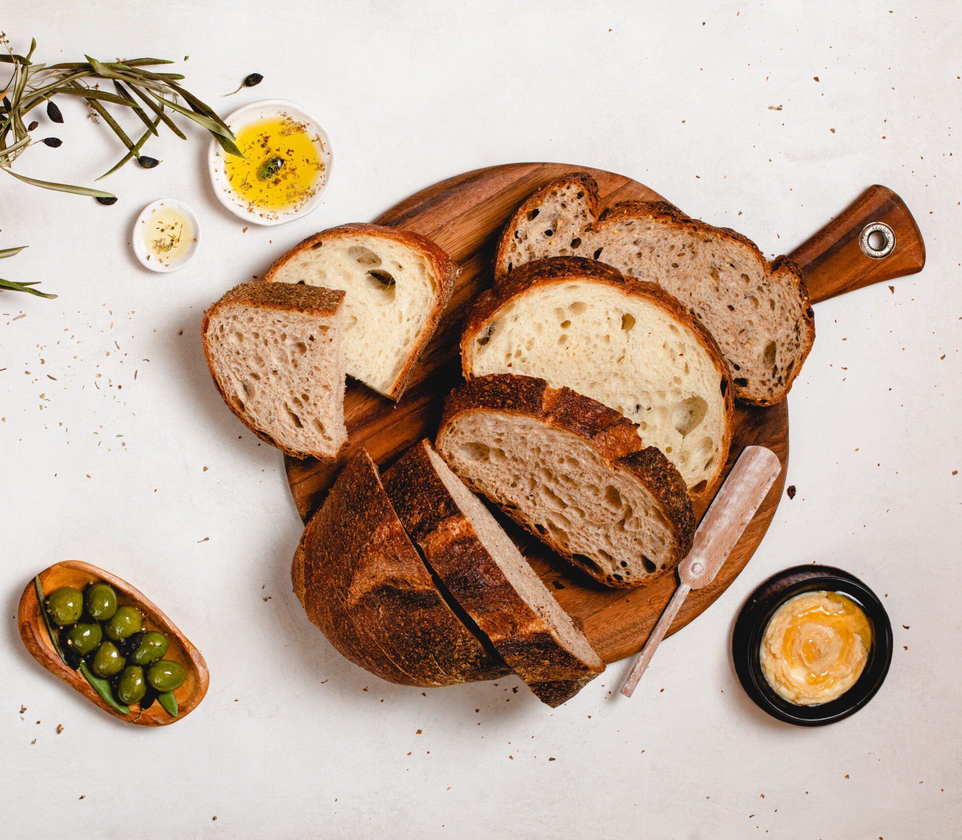 sliced loaf of bread on cutting board with herbs, olives, knife and melted butter